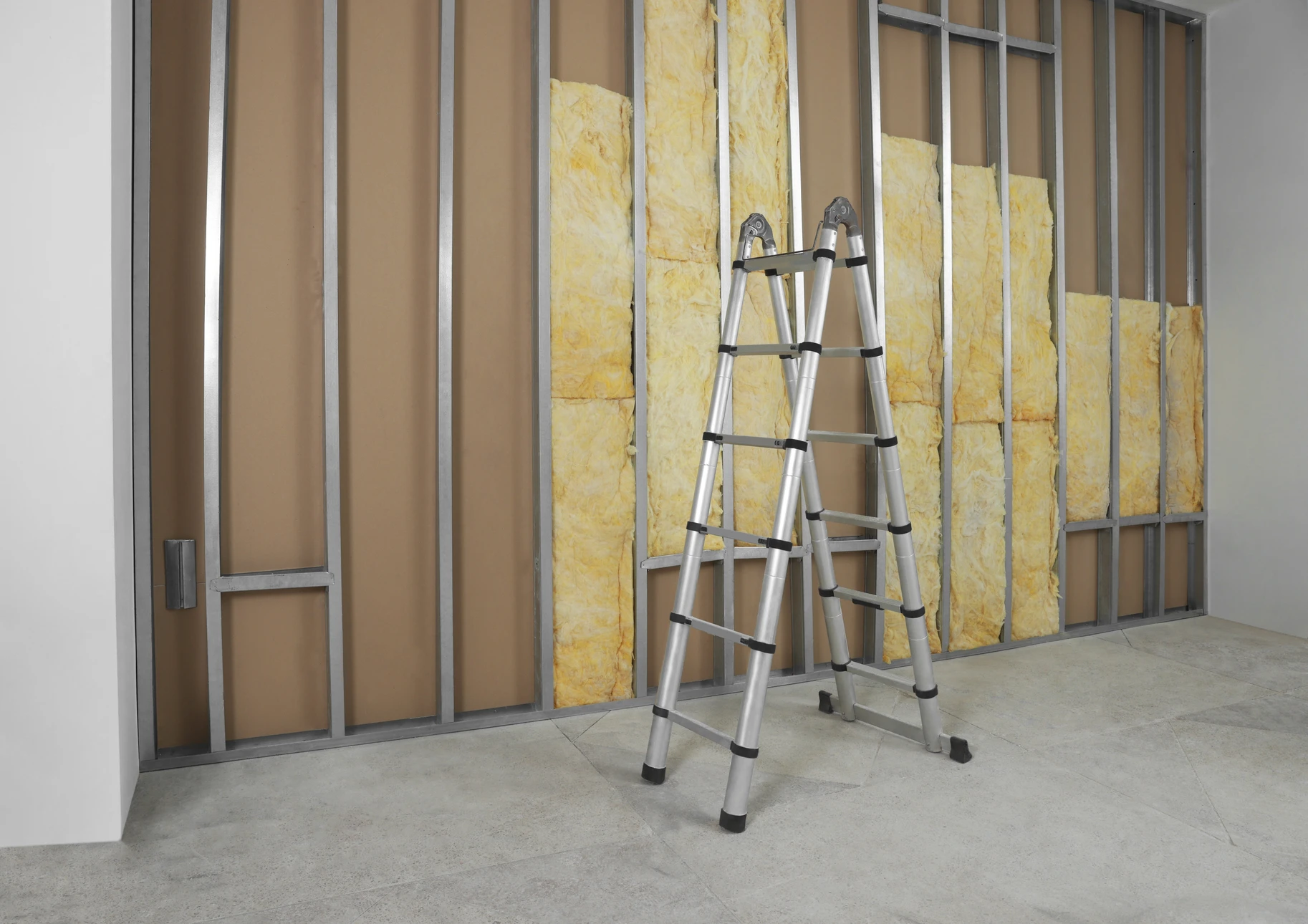 Intérieur de bâtiment en construction, ouvrier en combinaison de protection blanche et gants installant de la laine de verre jaune entre montants métalliques d'une cloison